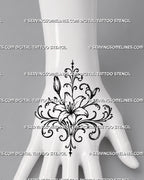 Ornamental lily tattoo shown across the back of the hand with wide mirrored filigree, a central flower, botanical buds, and wrist-facing details