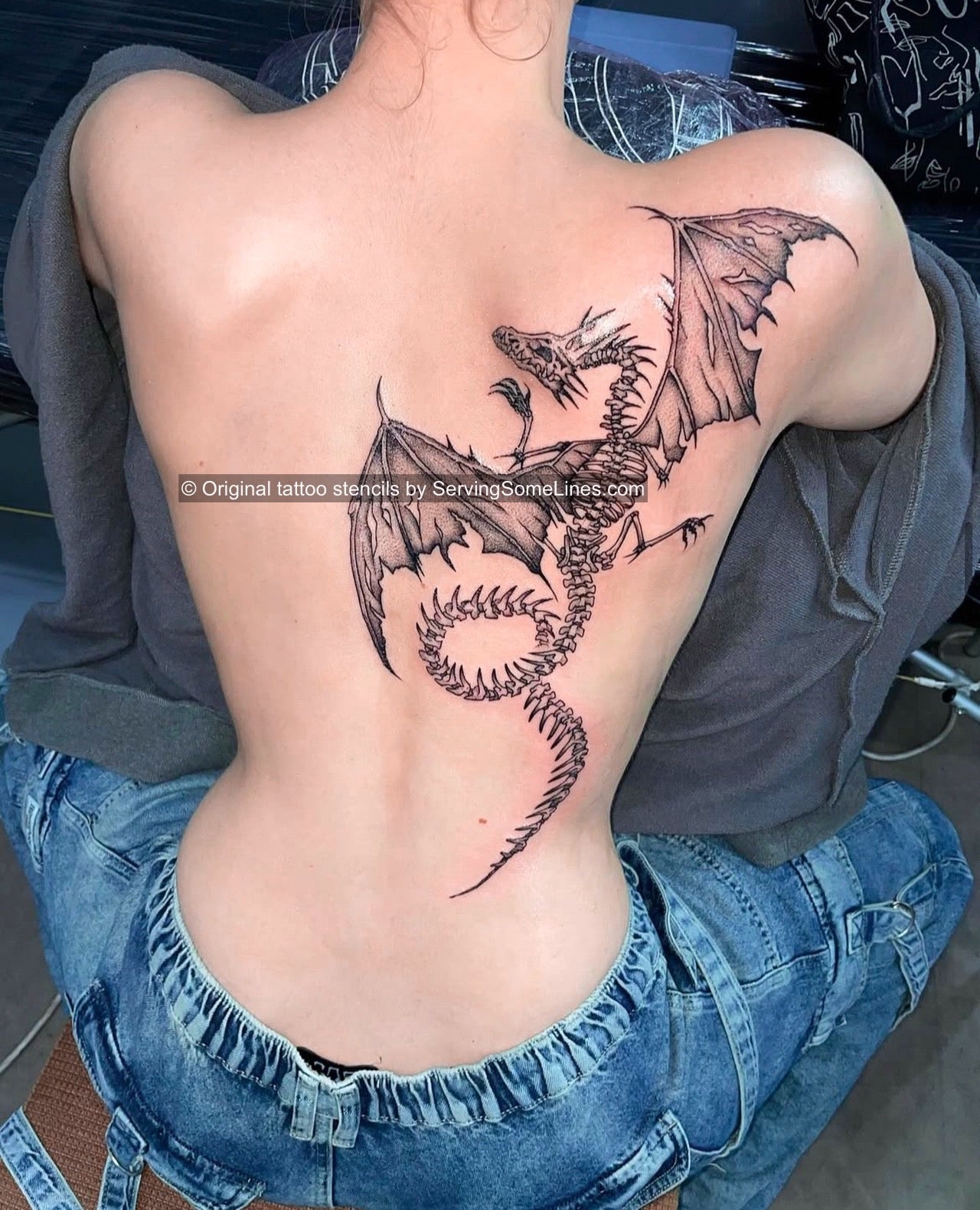 Winged dragon skeleton tattoo on girl’s back, detailed custom design with gothic wings and fine black linework along shoulder and spine
